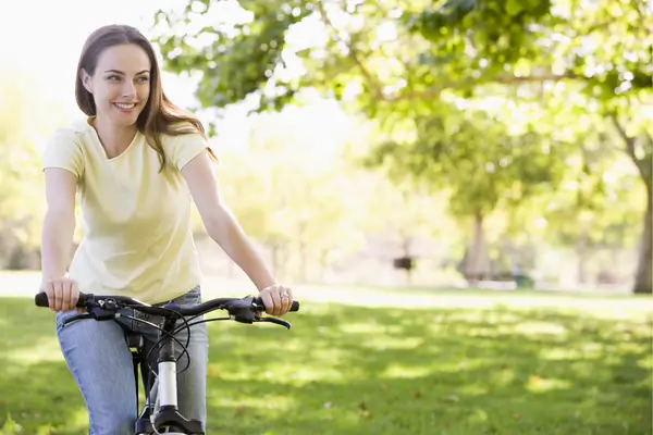 Mulher sorrindo ao pedalar bicicleta em parque ensolarado como ritual de autocuidado e mente leve.